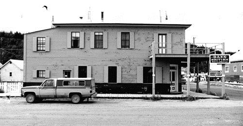 Side view of the Bank of British North America, showing details such as the ornate, Victorian-style front doors set into the salient angle, and the overhanging corner balcony supported on slender, turned porch columns, 1988. Photo by J. Mattie, via Parks Canada.
