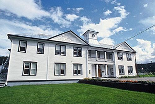 Front facade of the Former Territorial Court House in Dawson, showing its symmetrically organized multi-bay façade with a central entry, slightly projecting pedimented flanking pavilions, and the second-storey balcony with columns and balustrade, 1995. Photo by W. Lynch, via Parks Canada.