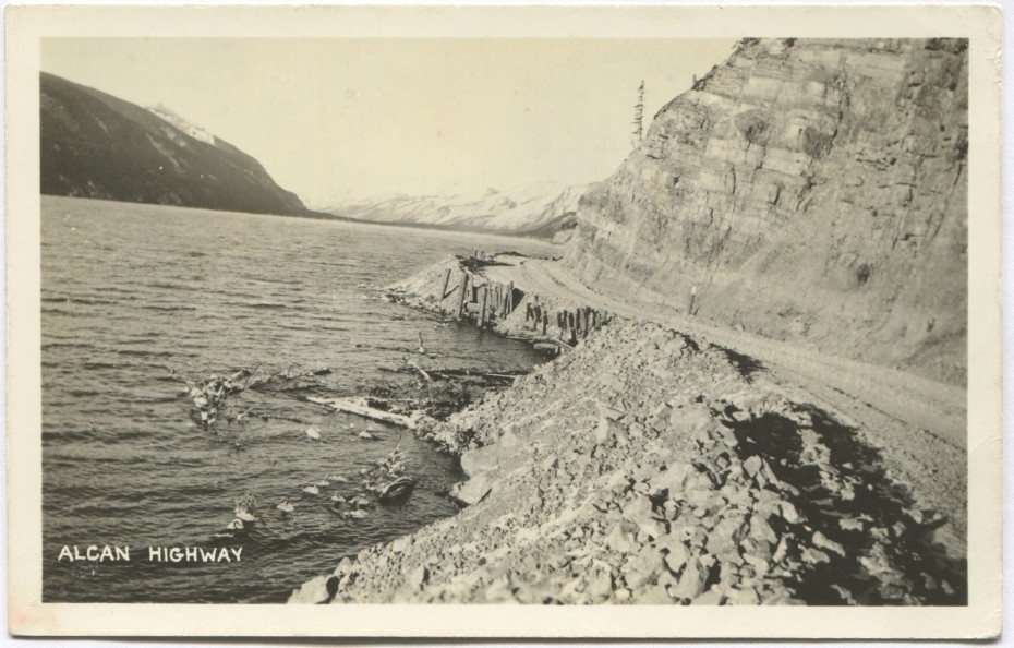 Postcard showing the gravel Alaska Highway along Muncho Lake.